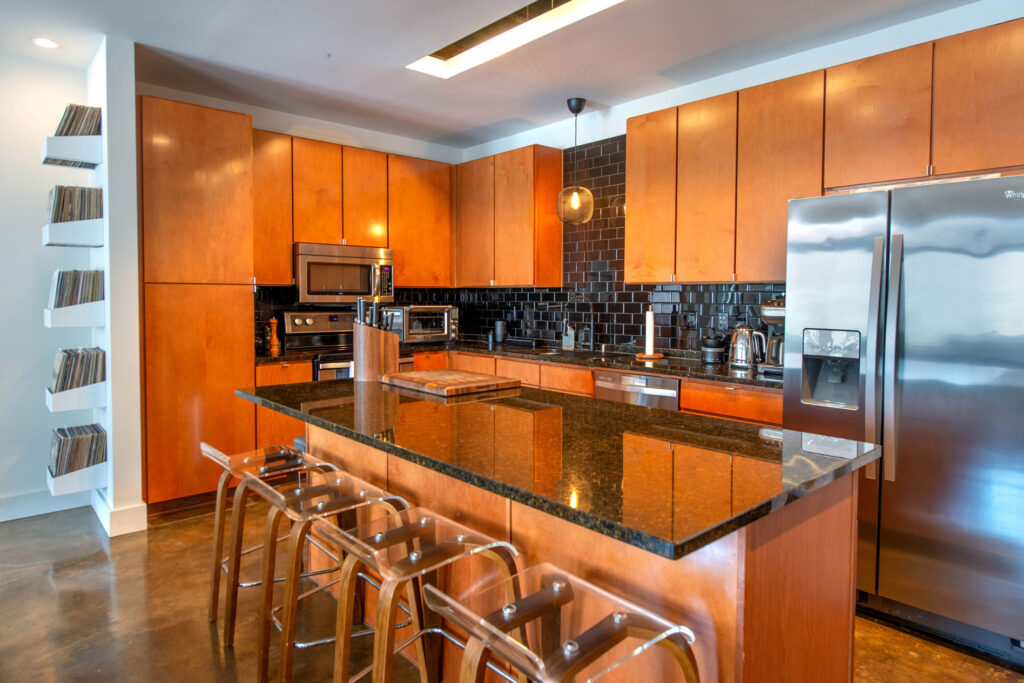 Modern kitchen with wooden cabinetry, a dark granite island, and four clear molded stools, featuring a wall-mounted record storage unit to the left.