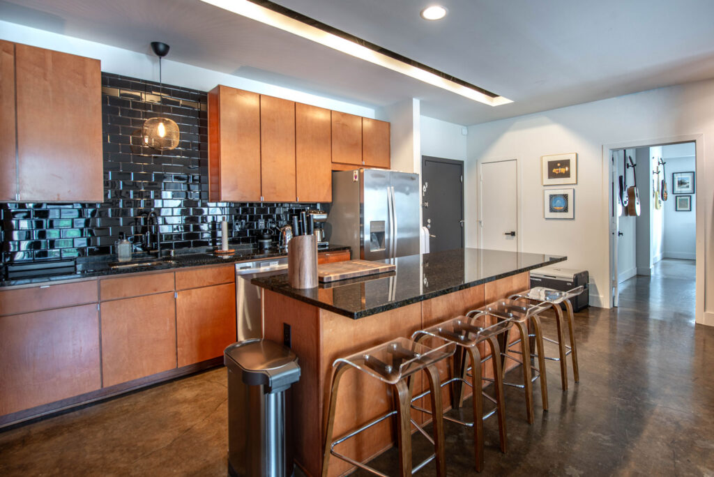 A modern kitchen with wood cabinets, black tile backsplash, and a black granite-topped island with four clear acrylic stools.