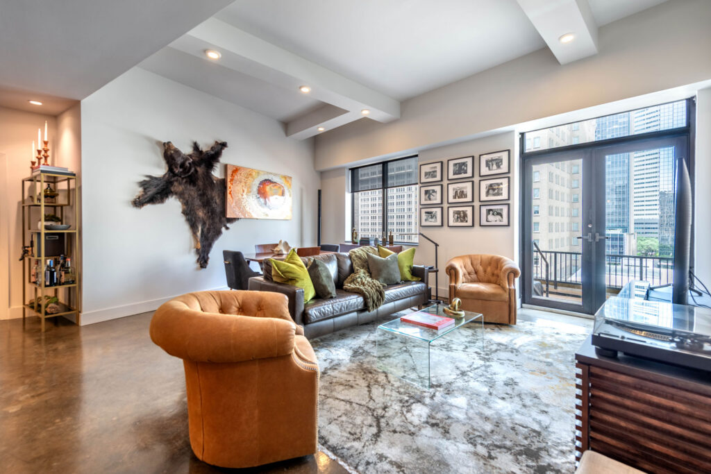A modern living room with a dark leather sofa, two tan armchairs, a glass coffee table, and a large bear pelt mounted on the wall.