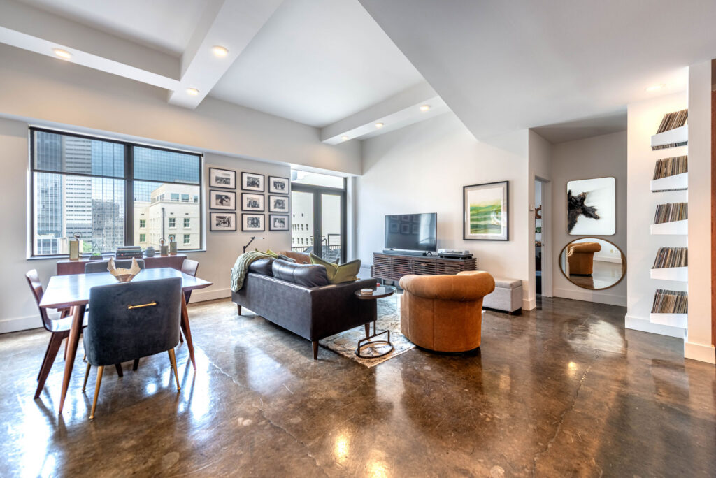 An open-concept living and dining area featuring a dark sofa, a tan armchair, a television, and a wall of framed photographs, with large windows overlooking a city skyline.