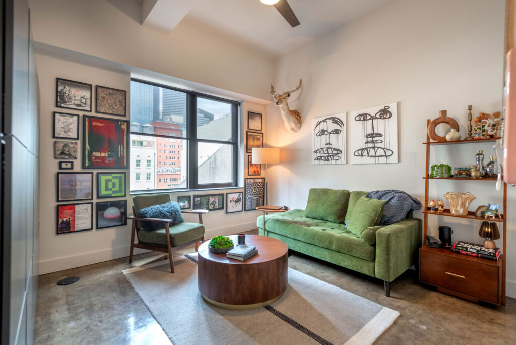 A living room featuring a green sofa, a green armchair, and a circular wooden coffee table, with a gallery wall of posters and artwork surrounding a window overlooking city buildings.