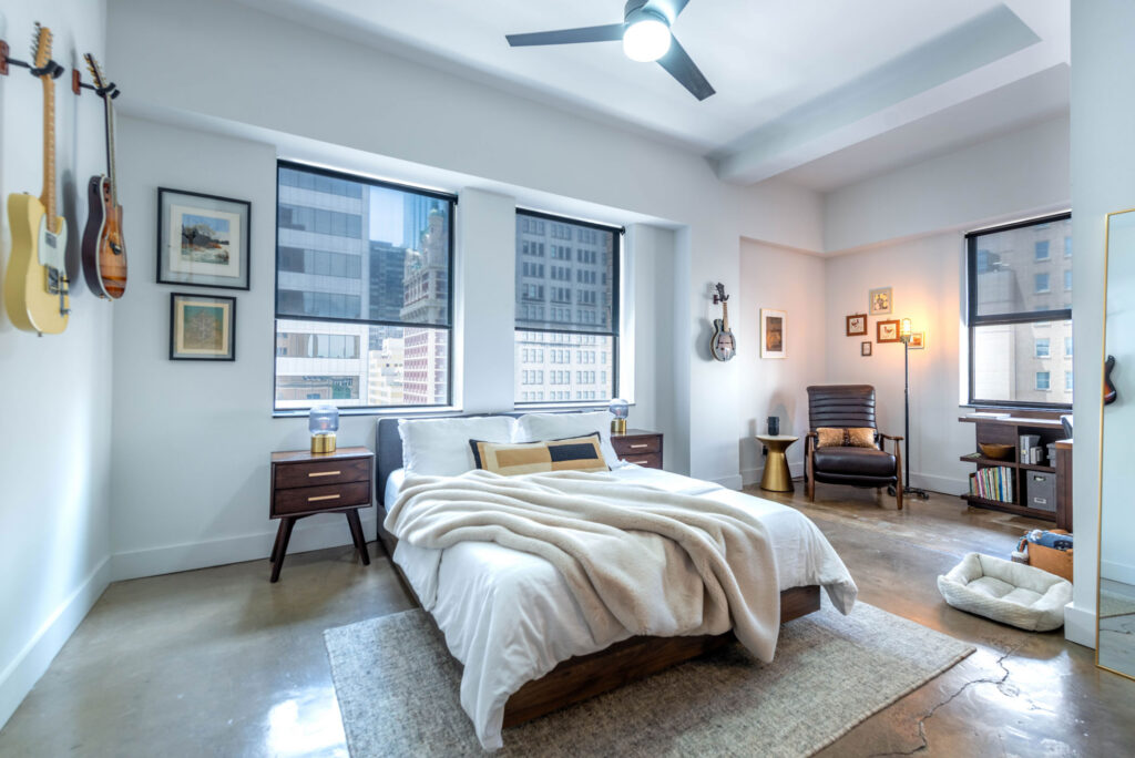 A bright bedroom featuring a queen bed with a white duvet, two nightstands, a leather armchair in the corner, and several acoustic and electric guitars hung on the walls.