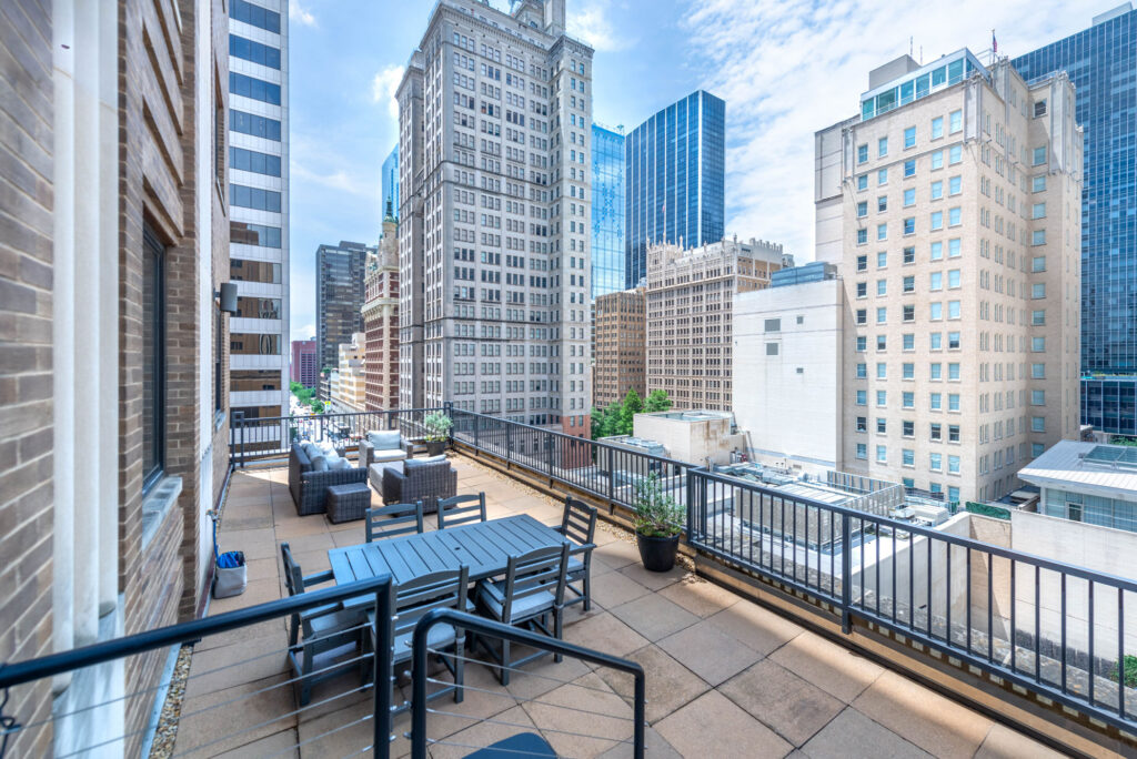 Rooftop terrace with a dining table and lounge seating, overlooking a dense city skyline of tall office buildings.