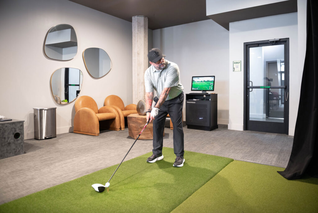 A man in a polo shirt and baseball cap swings a golf club on an indoor putting green in front of a golf simulator.