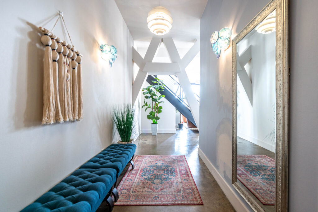 A hallway with a blue tufted bench, a patterned area rug, a potted plant, and a large wall mirror, leading to a staircase with white structural supports.