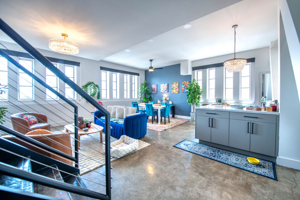 Open-plan living room with a blue sofa, dining area, and kitchen island featuring grey cabinets, viewed from the bottom of a staircase.