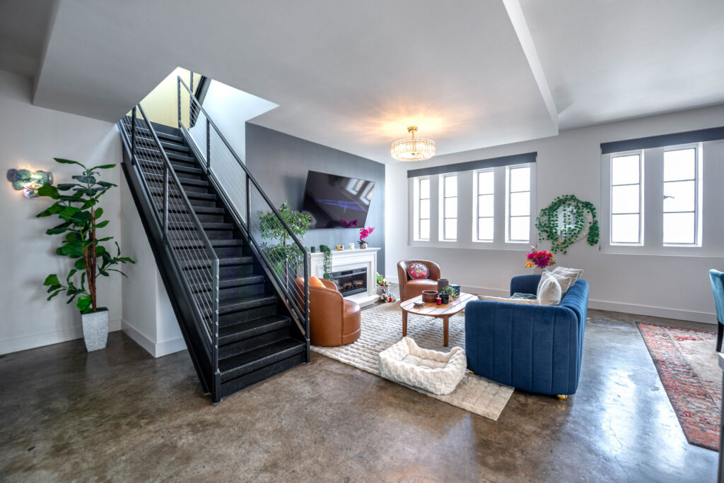 A living room with a concrete floor, a black staircase, a television above a fireplace, a blue sofa, and two brown armchairs on a rug.