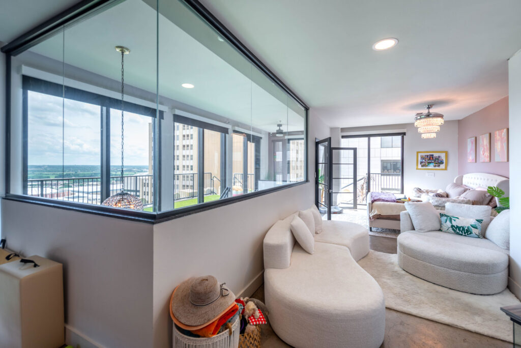 A modern living space featuring a curved white sofa, a glass-walled sunroom overlooking a city balcony, and an adjacent bedroom area.