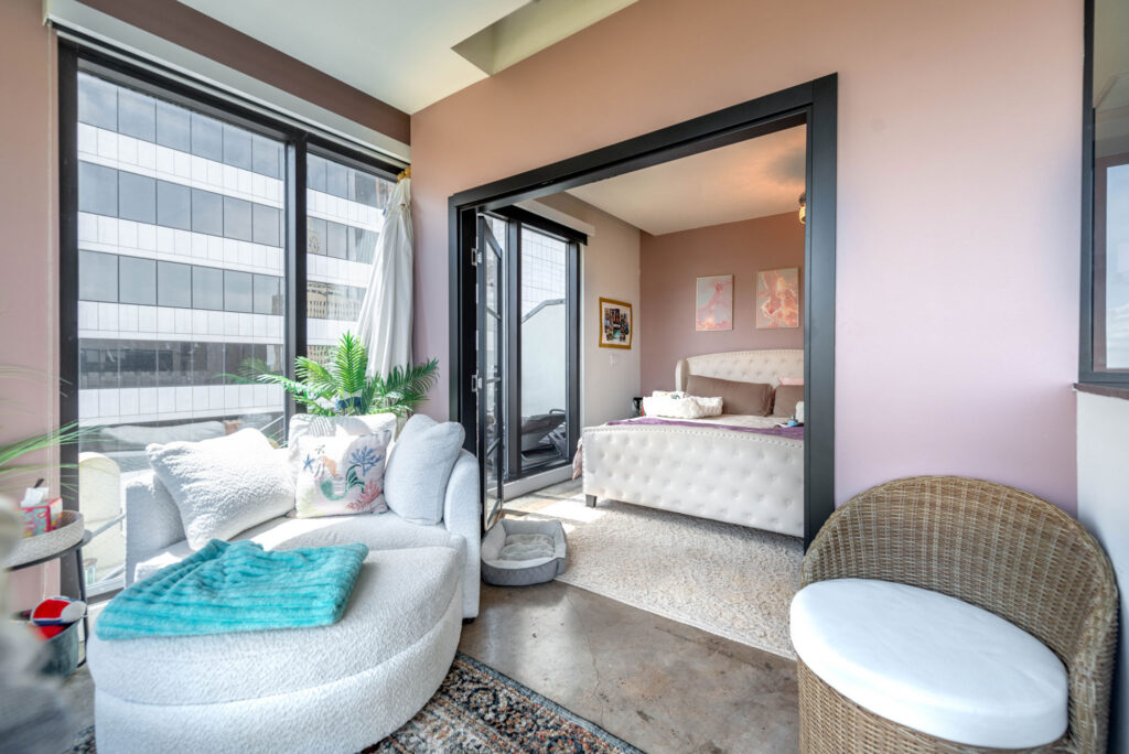 A living area with a white circular sofa and turquoise throw leads into a bedroom through a doorway, featuring a tufted bed, pink walls, and a view of a city building through large windows.