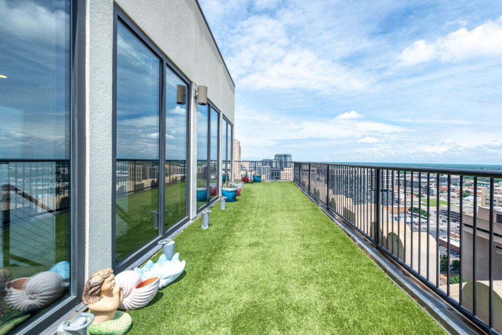 A balcony with artificial turf, lined with decorative pots and figurines against a floor-to-ceiling glass wall, overlooking a city skyline.